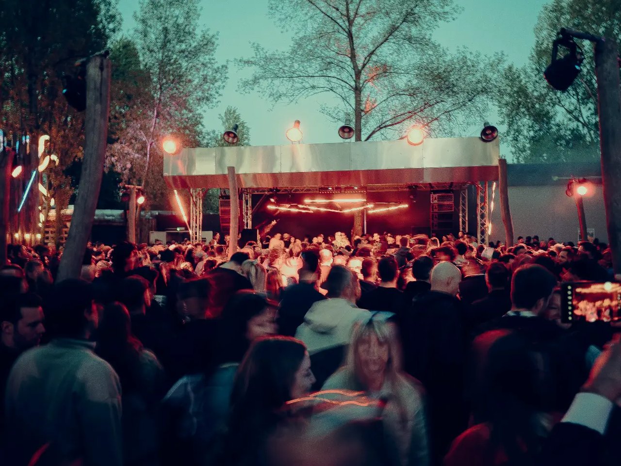 Große Menschenmenge vor einer beleuchteten Outdoor-Bühne im Westhafen Leipzig am Abend; warmes, rötliches Licht, Bäume im Hintergrund und eine dichte Festival- oder Konzertatmosphäre.